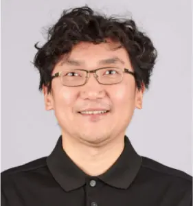 A person with curly hair and eyeglasses is smiling, wearing a black collared shirt against a plain background.