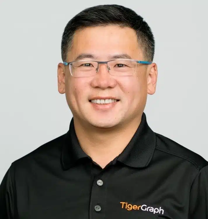 A person with short dark hair and glasses smiles at the camera. They are wearing a black polo shirt with the TigerGraph logo on it. The background is plain and light-colored.