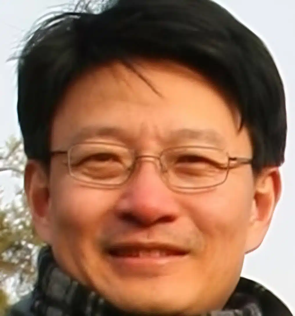 A man with short black hair and glasses is smiling. He is wearing a dark scarf and a jacket. The background is blurred.