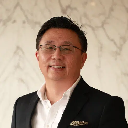 A person wearing glasses, a dark blazer, and a white shirt smiles at the camera. The background features a light-colored, marble-patterned wall.