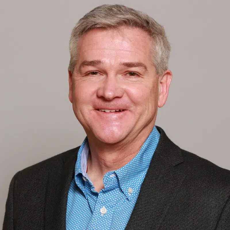 A smiling man with short gray hair, wearing a blue polka dot shirt and a dark blazer. The background is a plain light color.