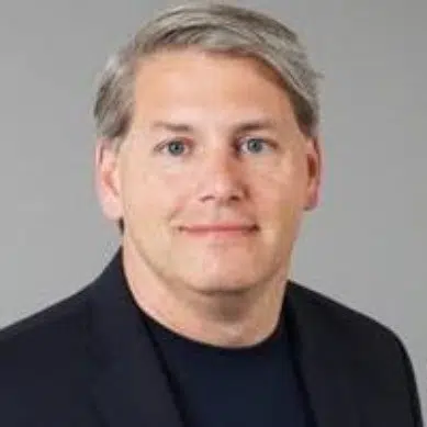 Portrait of a man with short gray hair, wearing a black blazer over a dark shirt. He is looking directly at the camera with a slight smile, set against a neutral gray background.