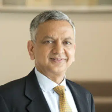 A middle-aged man with short gray hair, wearing a dark suit, light blue shirt, and yellow patterned tie, smiling slightly while looking at the camera, in a softly lit indoor setting.